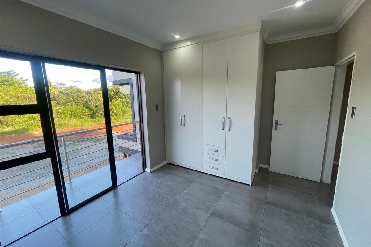 A modern, empty bedroom featuring sleek built-in cupboards, large format floor tiles, and a sliding glass door leading to a private balcony.