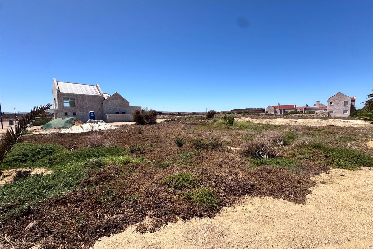 A wide-angle view of a vacant residential plot featuring natural coastal vegetation and an adjacent partially constructed stone dwelling.