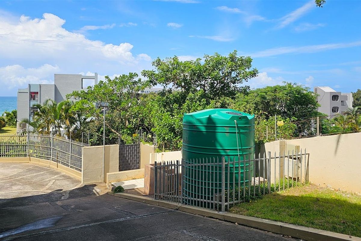 Exterior view of the apartment complex grounds featuring a large green water storage tank, perimeter fencing, and paved driveway access.