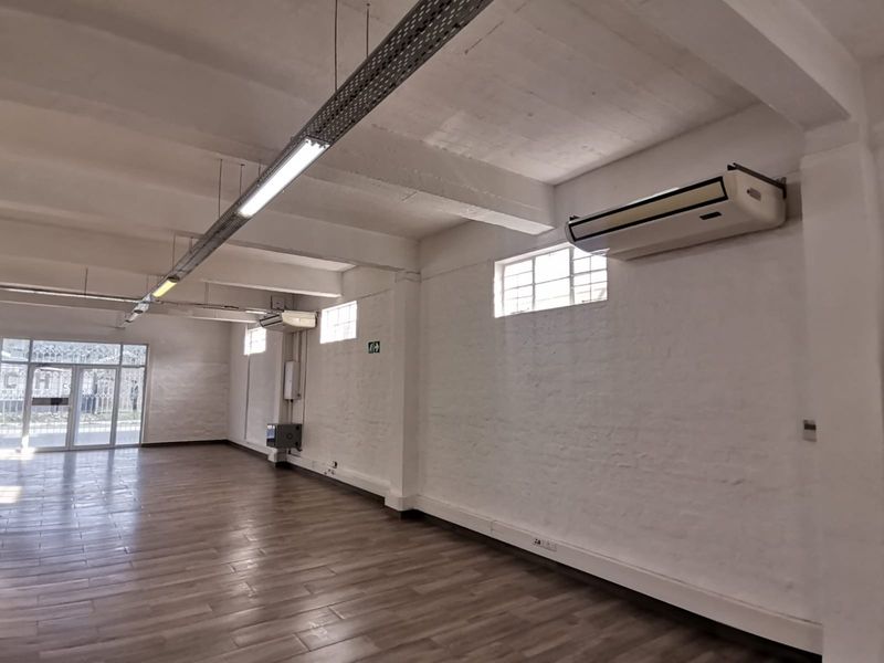 Spacious open-plan office area featuring high ceilings with exposed beams, modern tiled flooring, wall-mounted air conditioning, and ample natural light through high-level windows.