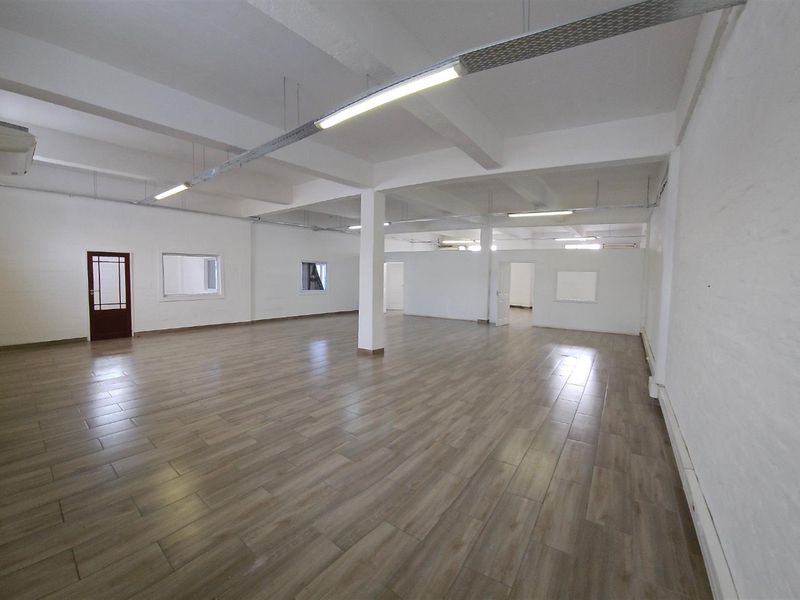 Spacious open-plan office area featuring wood-look tile flooring, exposed structural beams, and industrial-style overhead lighting.
