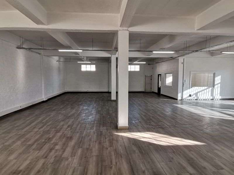 Spacious open-plan office area featuring exposed concrete beams, wood-look flooring, and ample natural light through multiple windows.