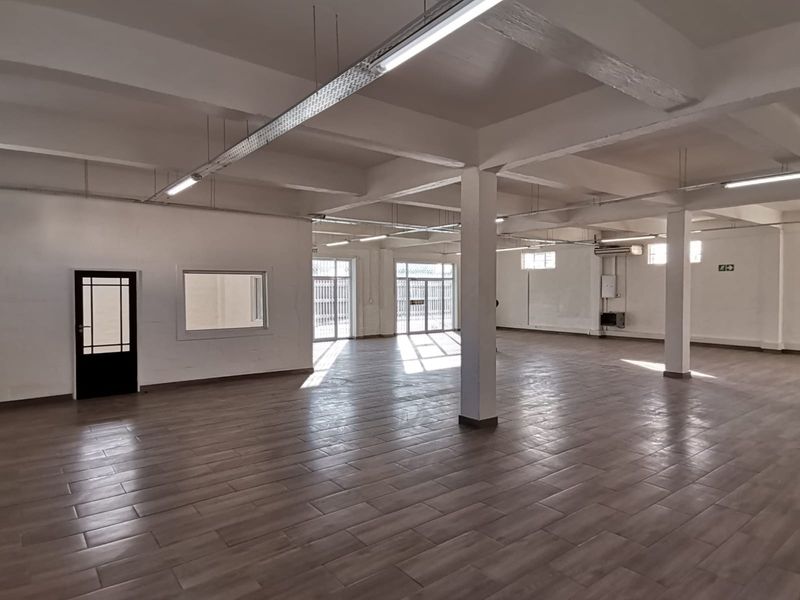 Spacious open-plan office area featuring tiled flooring, exposed structural beams, and ample natural light through multiple glass doors and windows.