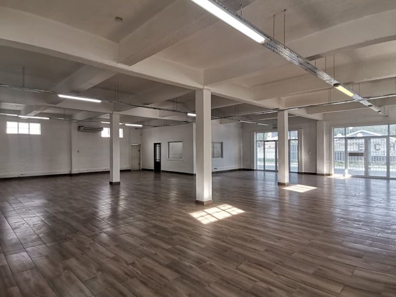 Spacious open-plan office area featuring wood-look tile flooring, exposed structural beams, and ample natural light through large glass entrance doors.