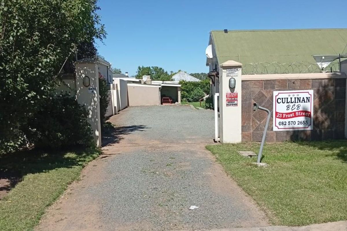 Street view of the guest house entrance featuring a gravel driveway, perimeter wall with security signage, and gated access.