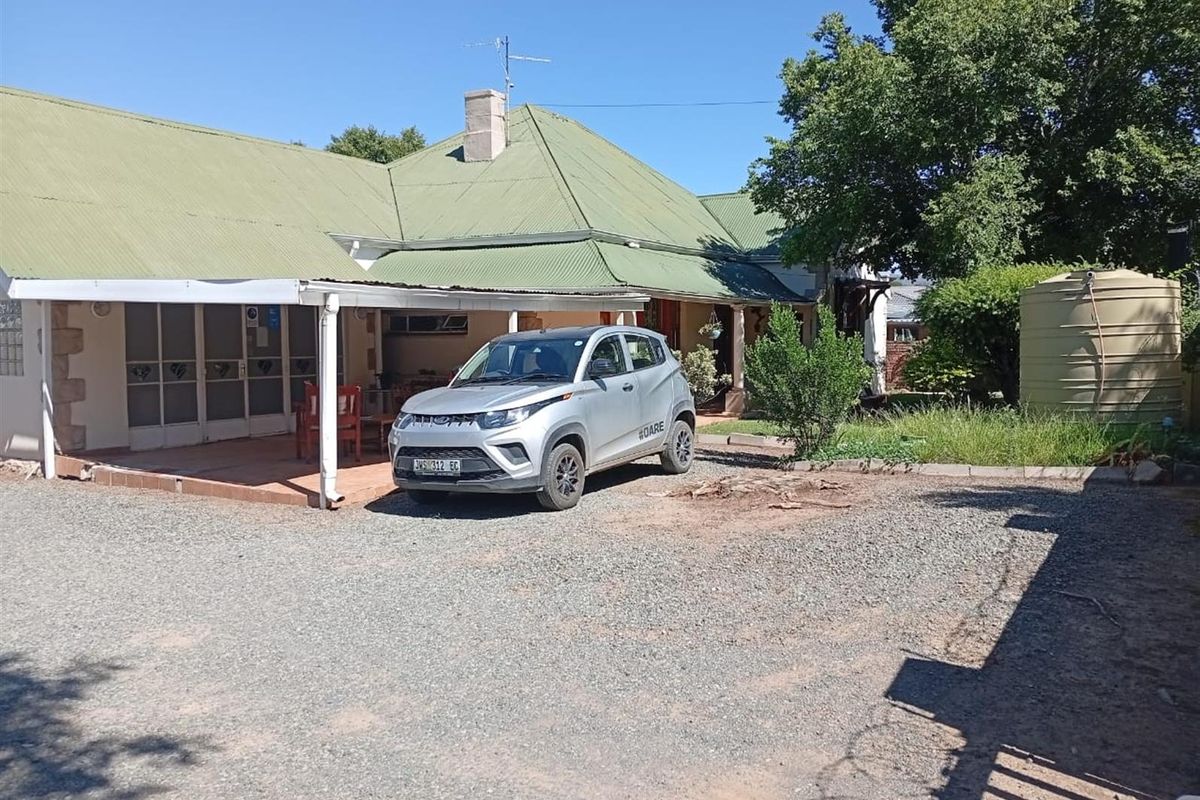 Exterior view of a guest house featuring a covered patio area, corrugated metal roof, and a gravel driveway with a water storage tank.