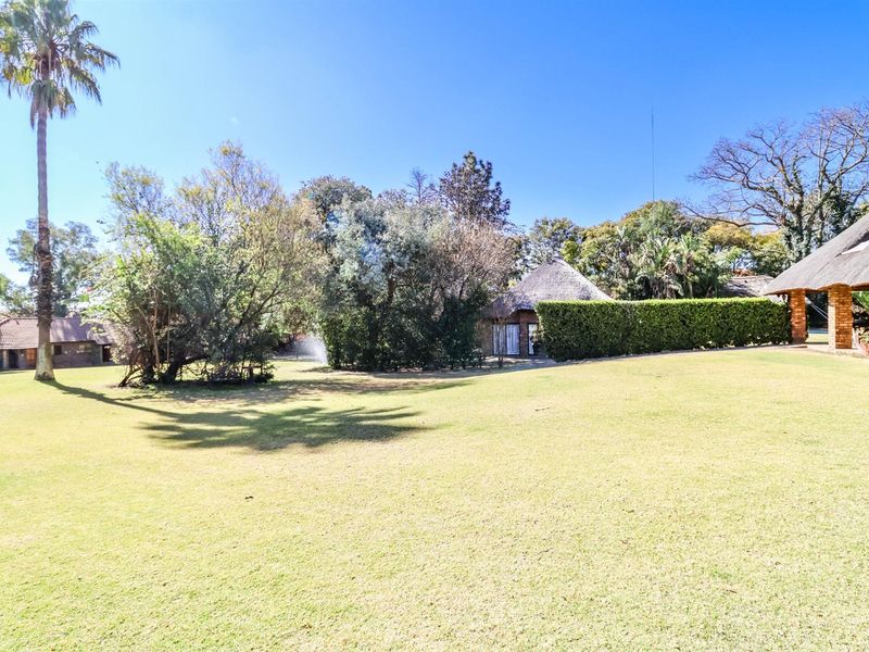 A spacious, well-maintained garden area featuring mature trees, manicured lawn, and glimpses of traditional thatched-roof structures on the property.