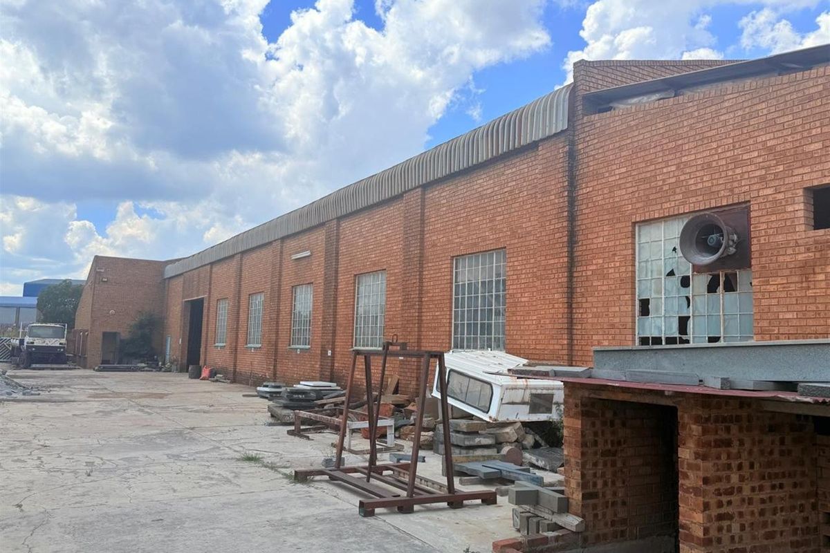 Exterior view of a commercial warehouse building featuring red brick facade, industrial windows, and concrete yard space requiring maintenance.