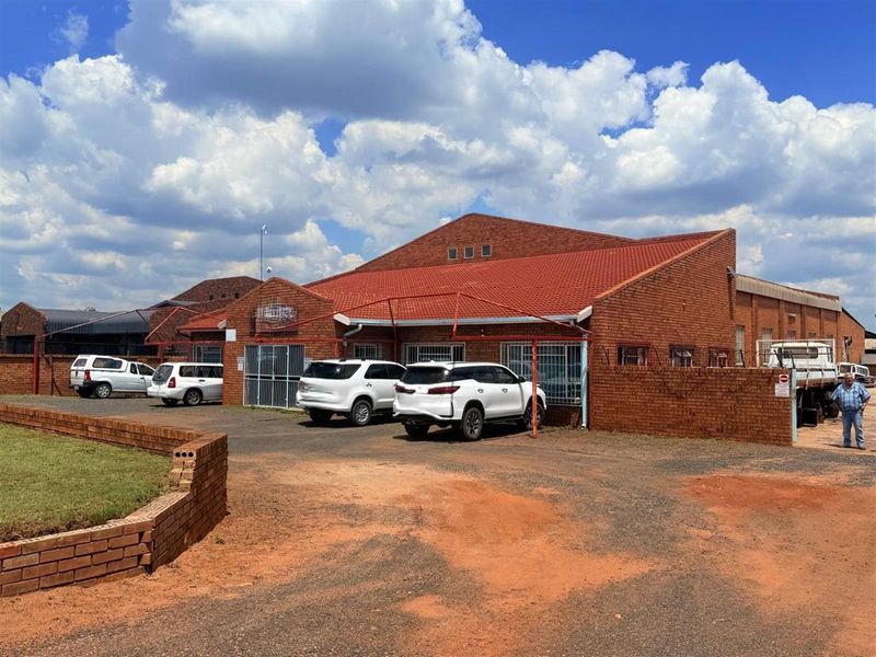 Exterior view of a commercial brick building featuring a pitched tiled roof, security gates, and an adjacent unpaved parking area.