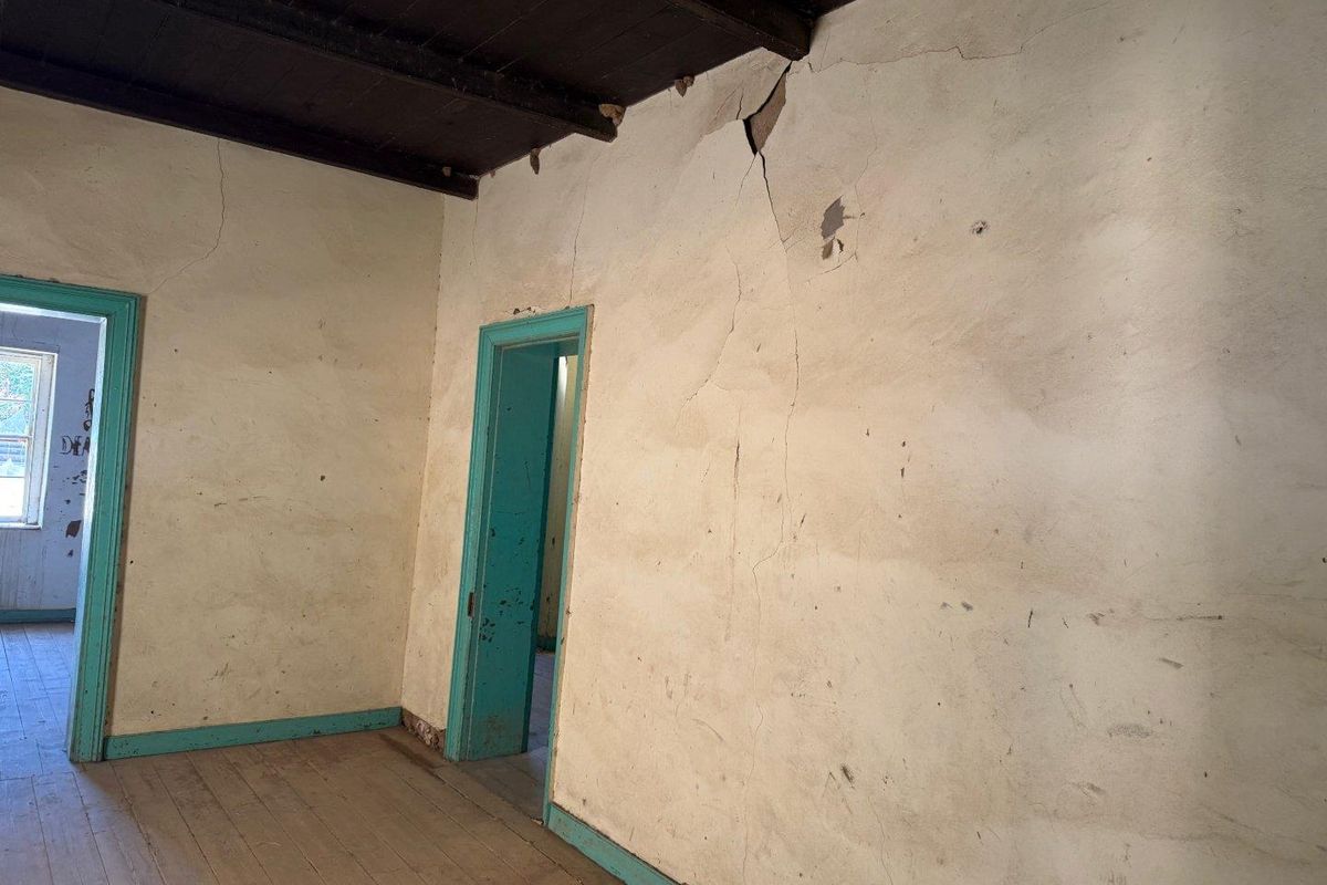 An interior room featuring exposed timber ceiling beams, original timber flooring, and significant wall plaster cracking requiring structural attention and cosmetic renovation.