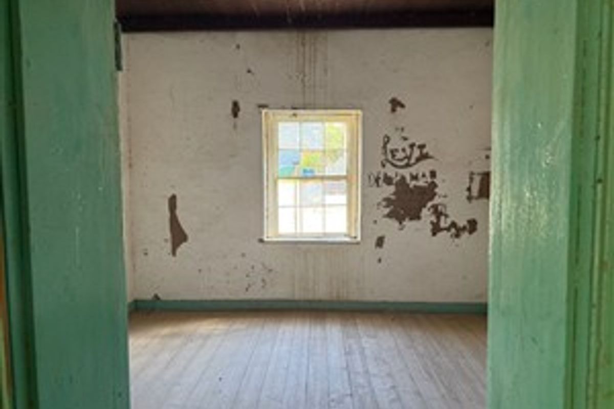 An empty bedroom featuring original timber flooring, exposed ceiling beams, and a sash window, requiring significant cosmetic restoration and wall repairs.