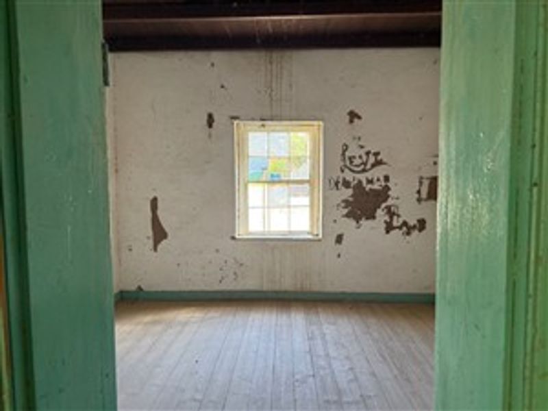 An empty bedroom featuring original timber flooring, exposed ceiling beams, and a sash window, requiring significant cosmetic restoration and wall repairs.