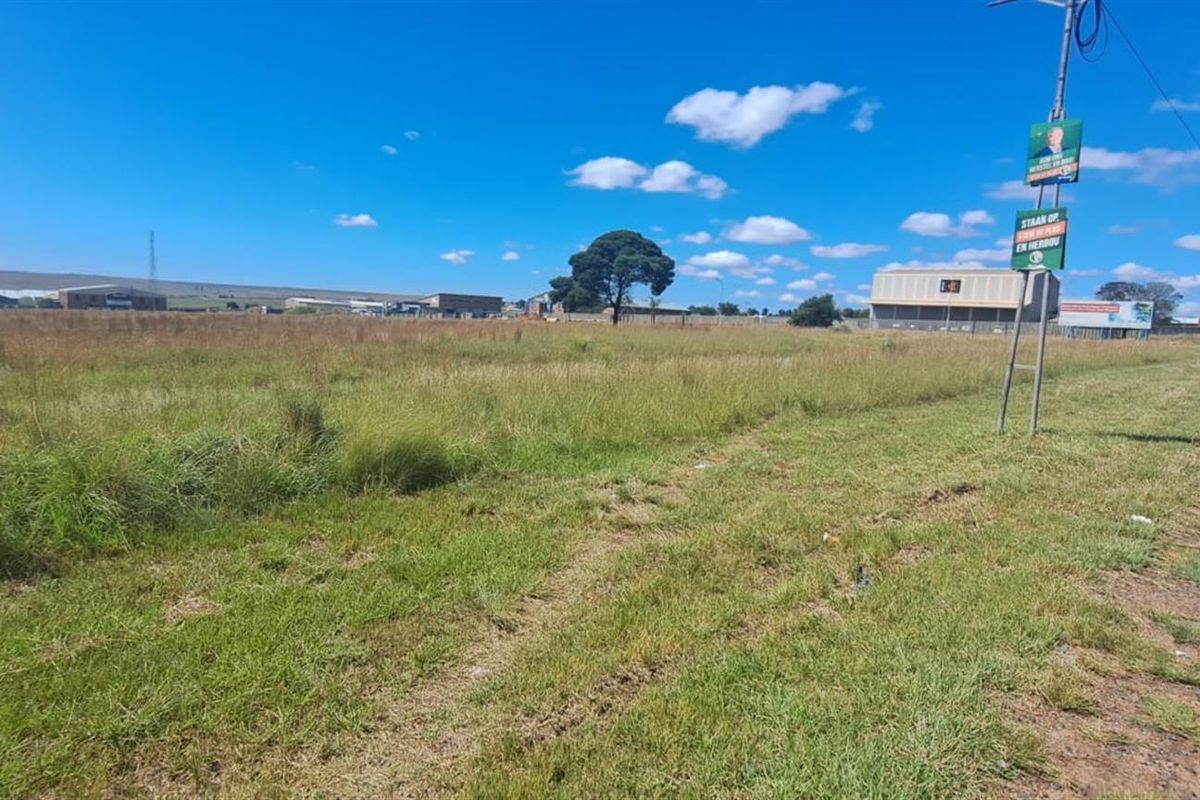 A wide-angle view of a vacant agricultural land plot situated adjacent to a public road, featuring open grass fields and surrounding industrial structures.