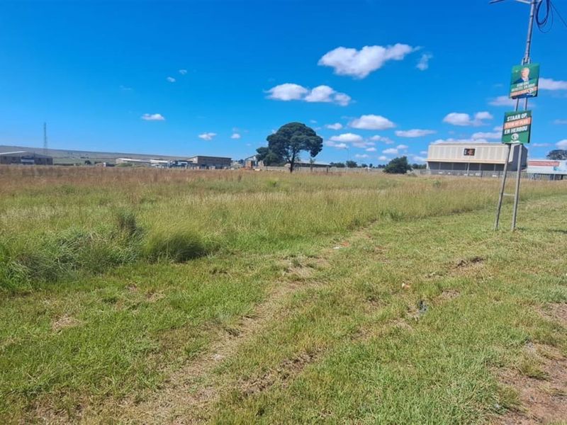 A wide-angle view of a vacant agricultural land plot situated adjacent to a public road, featuring open grass fields and surrounding industrial structures.