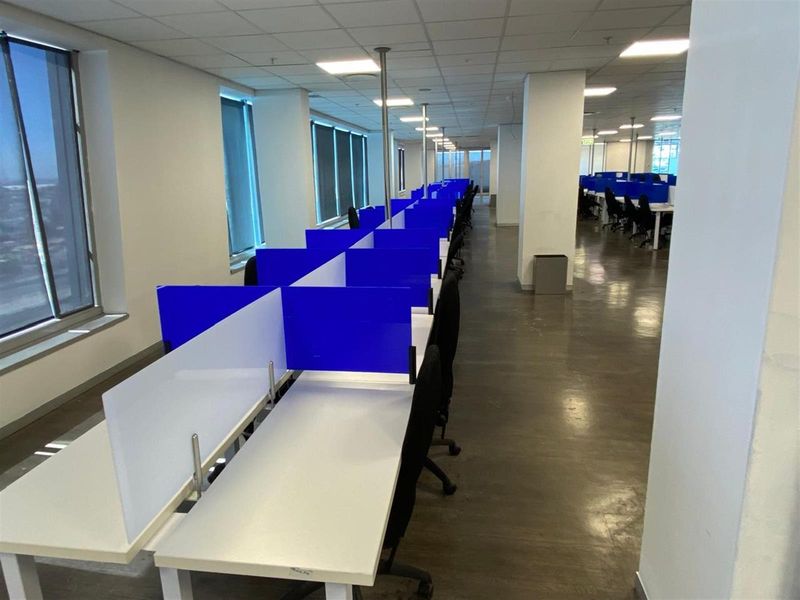 Open-plan office space featuring rows of built-in workstations with privacy screens, large windows, and suspended ceiling grid with integrated lighting.