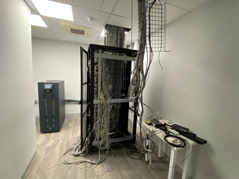 A dedicated server room featuring a central network rack with extensive exposed cabling, a floor-standing UPS unit, and suspended ceiling tiles.