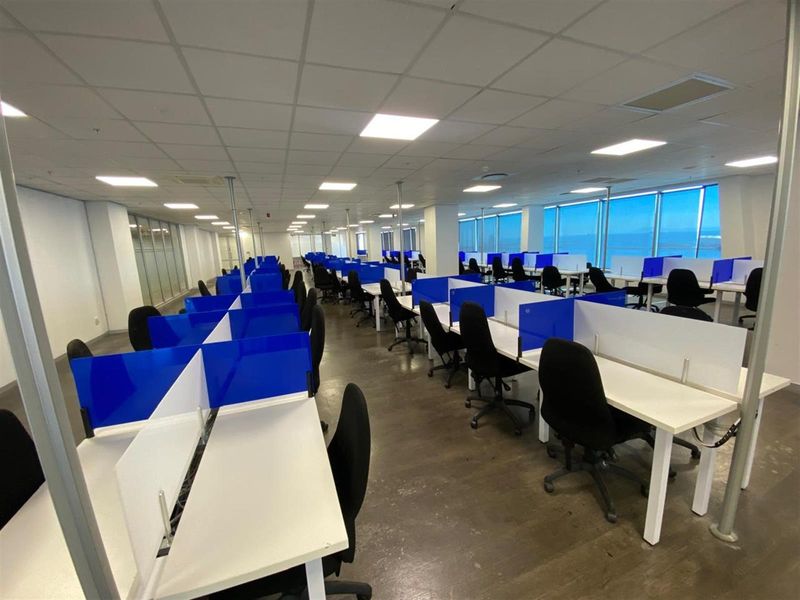 Spacious open-plan office area featuring rows of built-in workstations with privacy screens, suspended ceiling grid, and large windows offering natural light.