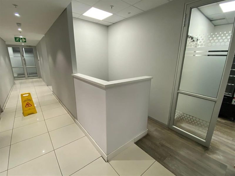 A bright office hallway featuring tiled flooring, recessed ceiling lighting, and a glass-partitioned office entrance with frosted privacy detailing.
