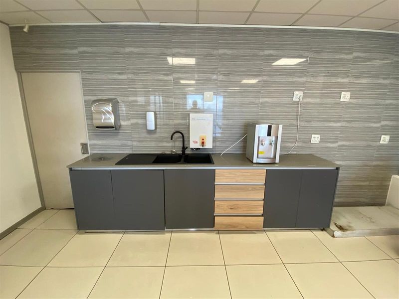 A functional office kitchenette featuring modern grey cabinetry, wood-look drawers, a black sink with a matte black faucet, and tiled splashback.
