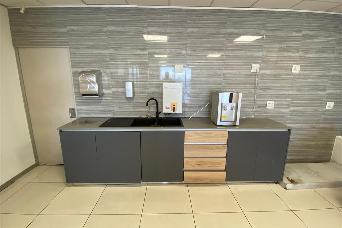 A functional office kitchenette featuring modern grey cabinetry, wood-look drawers, a black sink with a matte black faucet, and tiled splashback.