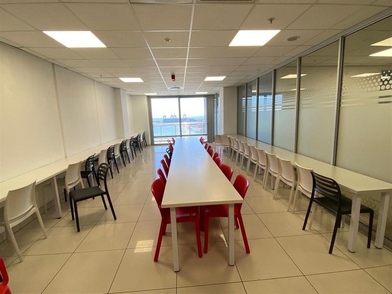 Spacious office area featuring a long central meeting table, perimeter desk space, tiled flooring, suspended ceiling, and large windows with coastal views.