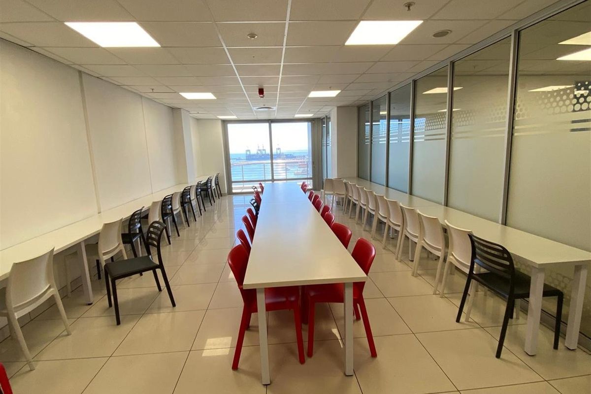 Spacious office area featuring a long central meeting table, perimeter desk space, tiled flooring, suspended ceiling, and large windows with coastal views.