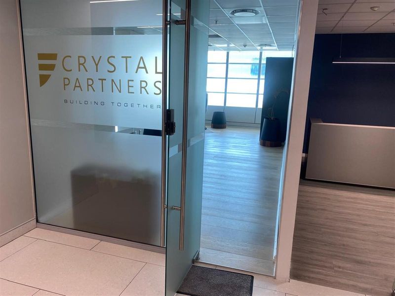 Modern office entrance featuring a glass door with branding, leading into a bright, open-plan workspace with suspended ceilings and laminate flooring.
