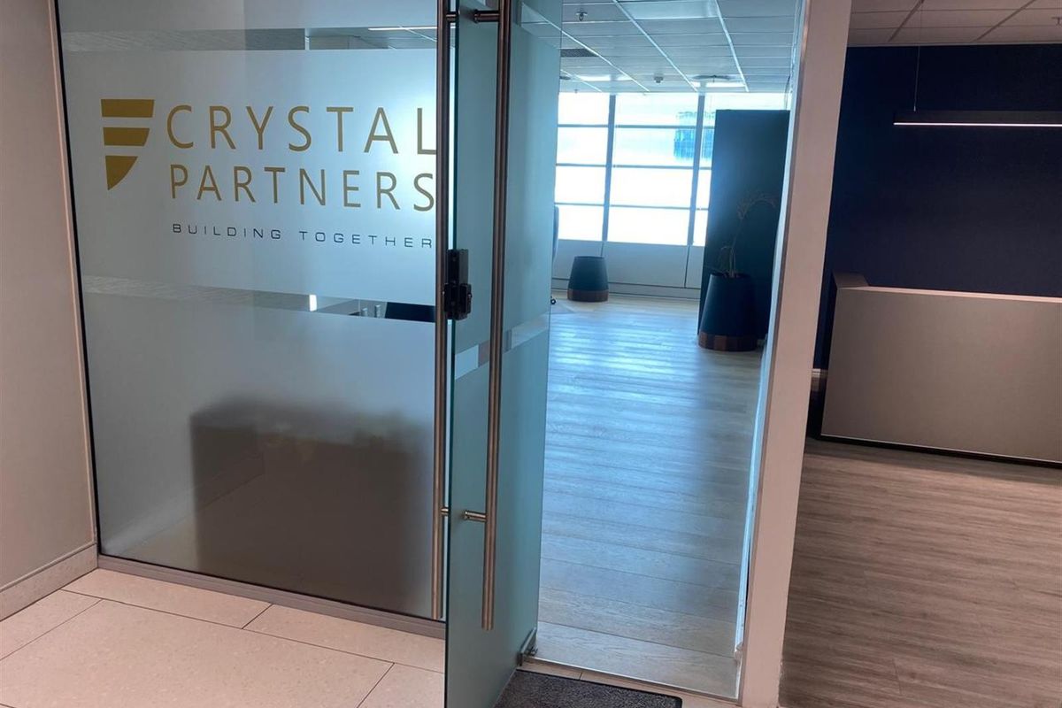 Modern office entrance featuring a glass door with branding, leading into a bright, open-plan workspace with suspended ceilings and laminate flooring.