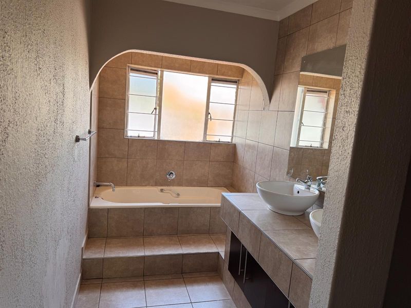 A functional bathroom featuring a built-in bathtub, tiled vanity with dual vessel sinks, and neutral wall tiling under a decorative archway.