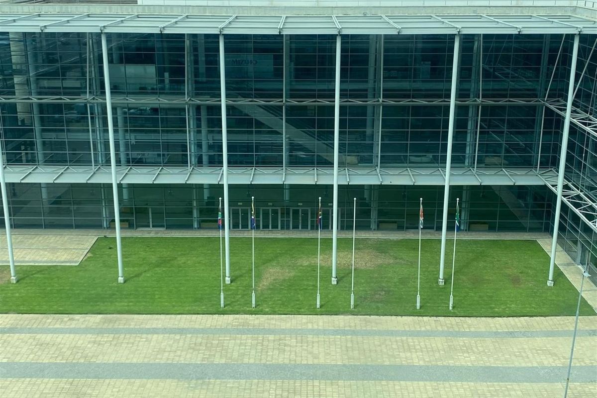 An elevated view of a modern office building facade featuring extensive glass curtain walling, structural steel supports, and a landscaped courtyard with flagpoles.
