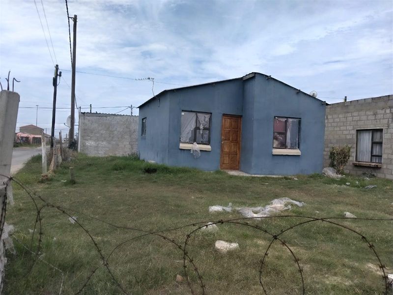 A detached single-story house featuring a blue painted exterior, timber entrance door, and metal-framed windows, situated on a grassed plot with boundary walling.