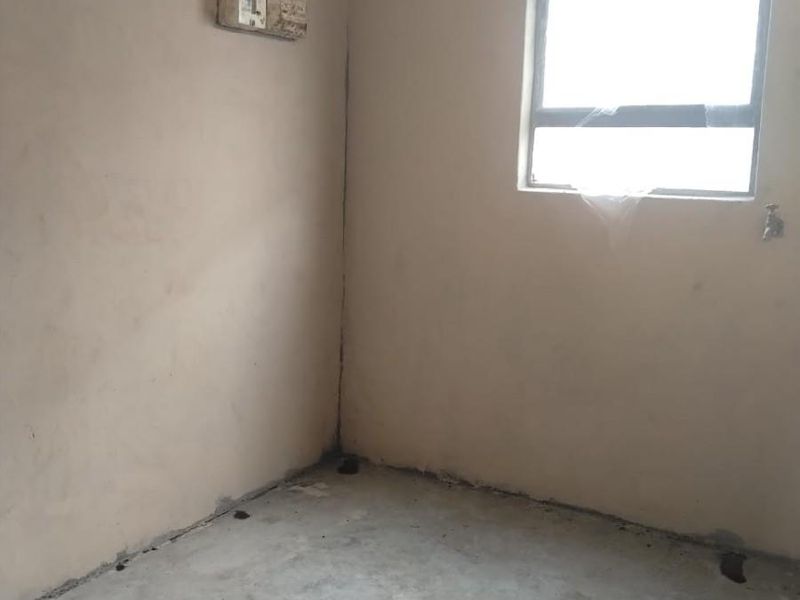 An unfinished interior space featuring raw concrete flooring, unpainted plastered walls, and a basic aluminium window frame with a wall-mounted prepaid electricity meter.