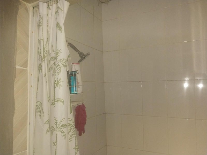 A functional shower enclosure featuring white ceramic wall tiling and a standard shower head, currently showing signs of wear and unfinished wall edges.