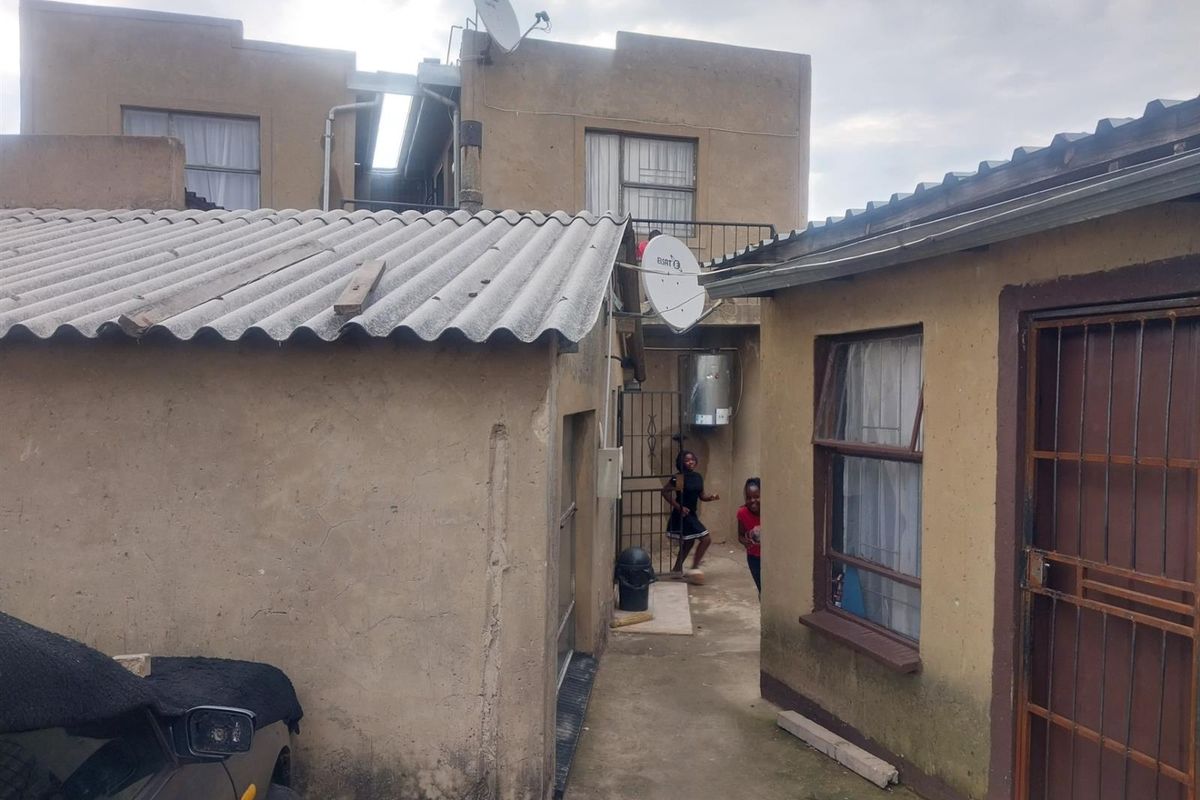 Rear exterior view of the property showing multiple structures with weathered plaster finishes, corrugated roofing, and security features.
