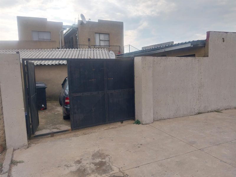 Front exterior view showing a metal driveway gate, concrete boundary wall, and paved driveway area leading to the property entrance.
