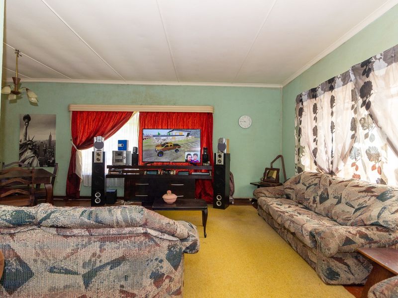 A spacious living room featuring carpeted flooring and large windows with curtain rails, requiring cosmetic updates to the wall finishes and flooring.