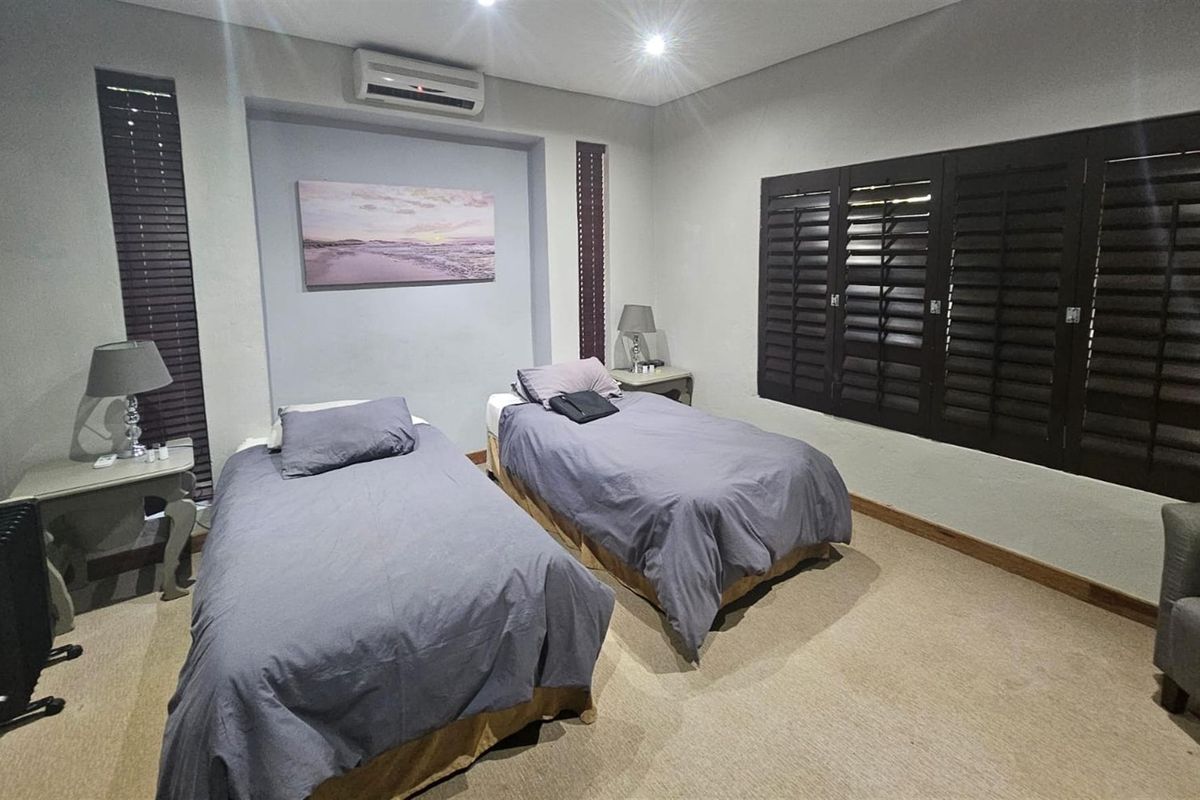 A well-maintained bedroom featuring neutral carpeting, recessed lighting, a wall-mounted air conditioning unit, and dark wooden plantation shutters.