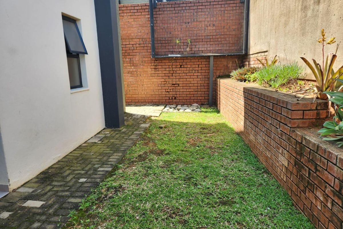 A compact rear garden area featuring a paved walkway, a small lawn section, and tiered brick retaining walls with established garden beds.