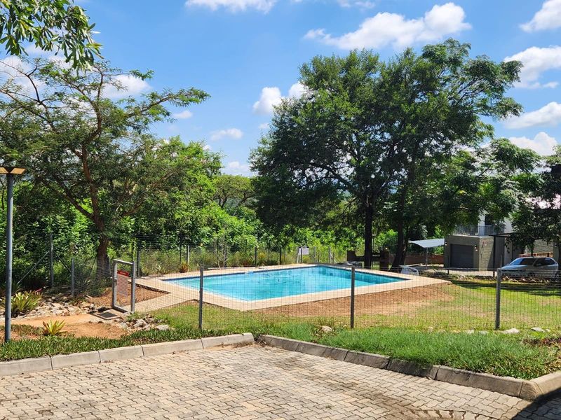 A communal swimming pool area featuring a paved surround, enclosed by wire fencing, set within a landscaped garden environment.