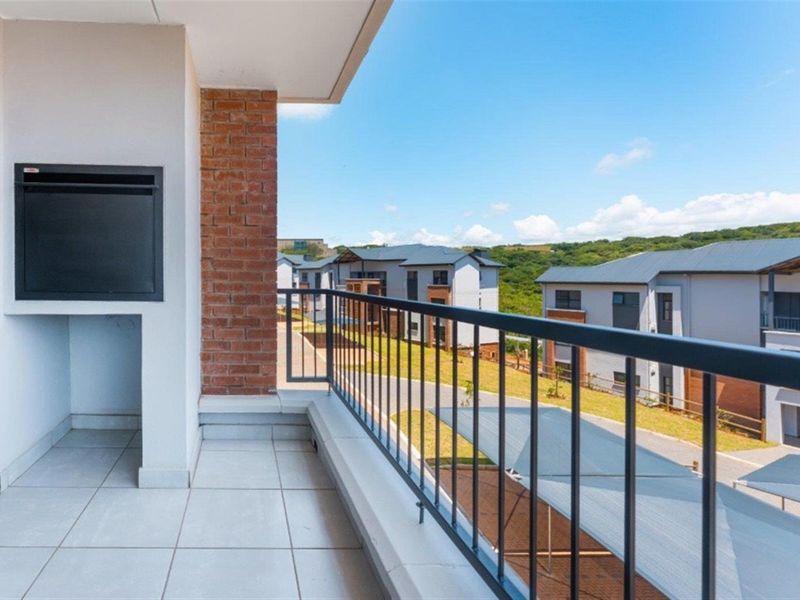 A clean, modern balcony featuring a built-in braai, tiled flooring, and a metal balustrade, offering pleasant views of the surrounding estate.