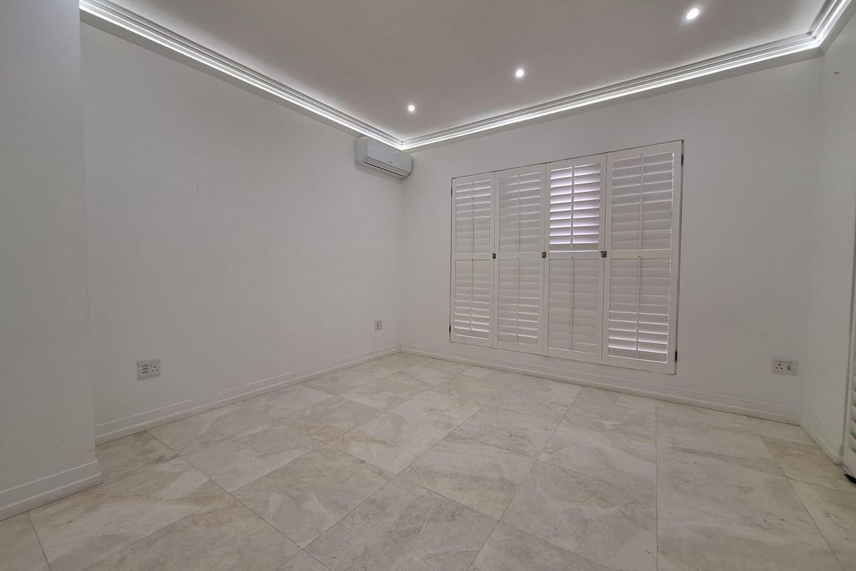 A bright, empty bedroom featuring neutral tiled flooring, recessed ceiling lighting with cove detail, white plantation shutters, and a wall-mounted air conditioning unit.