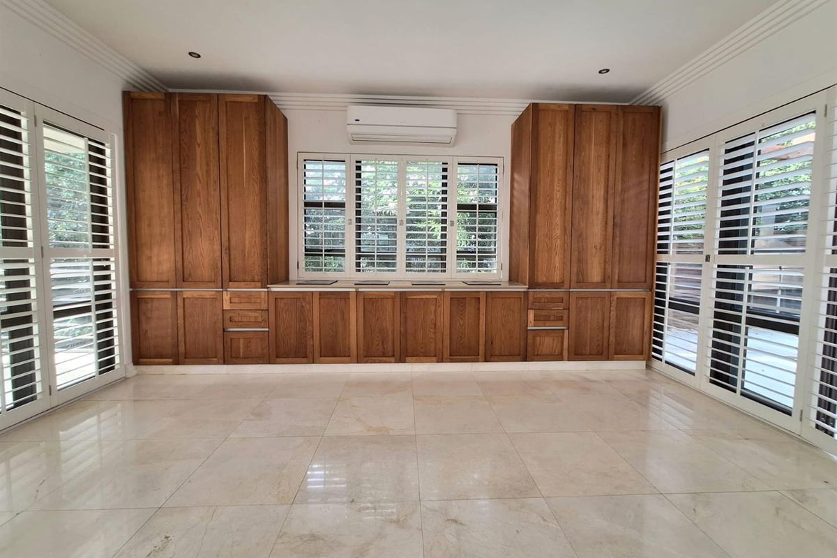 Spacious living area featuring custom built-in wooden cabinetry, large windows with plantation shutters, tiled flooring, and a wall-mounted air conditioning unit.