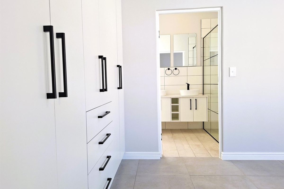 A modern bedroom featuring extensive built-in cabinetry with matte black hardware, leading into a contemporary en-suite bathroom with a glass-enclosed shower.