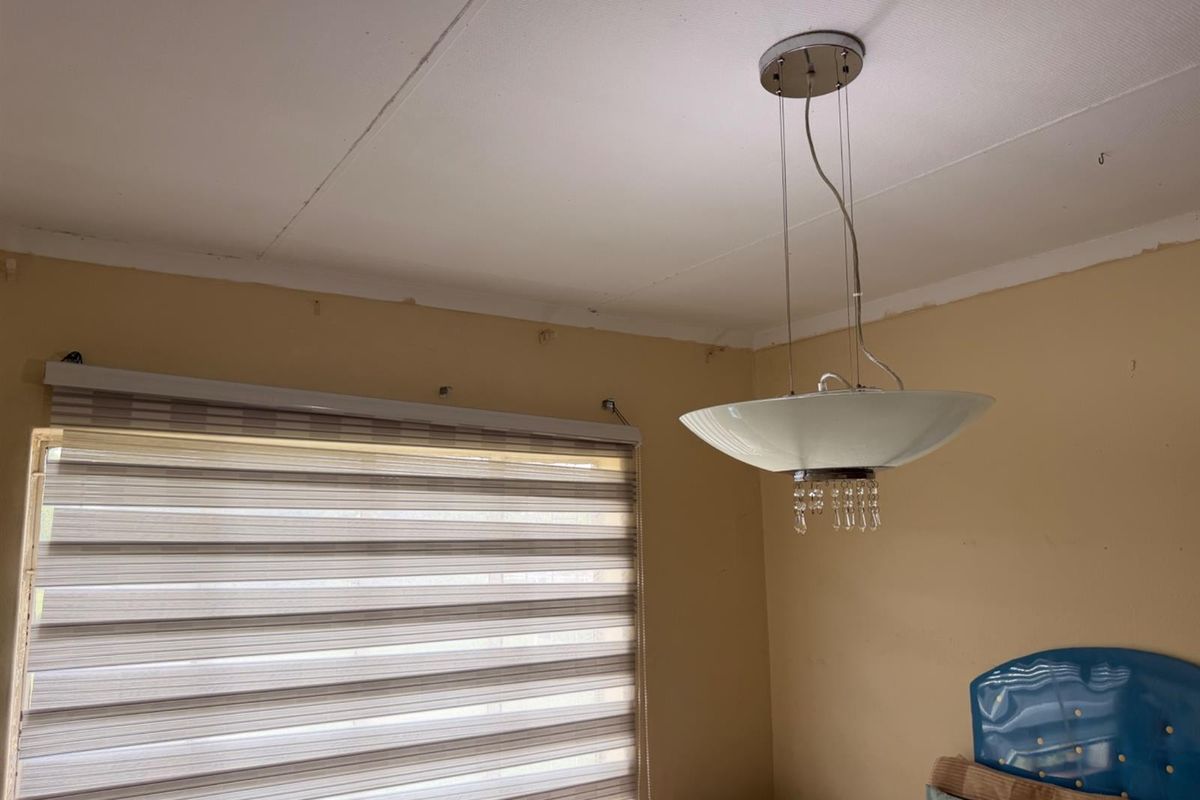 A bedroom interior featuring a modern pendant light fixture and fitted zebra-style roller blinds, showing signs of wear on the ceiling and walls.