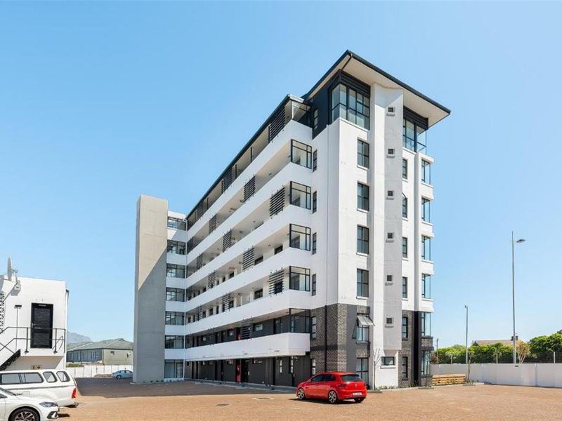 A modern multi-story apartment building featuring a clean white and dark facade with private balconies and an adjacent paved parking area.