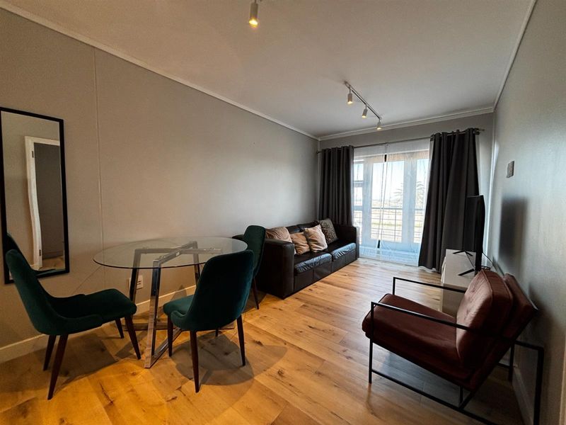 A well-lit living and dining area featuring timber flooring, modern track lighting, and direct access to a balcony through sliding glass doors.