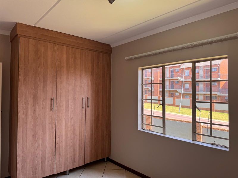 A bright, empty bedroom featuring wood-finish built-in wardrobes, tiled flooring, and a window with security bars overlooking the complex.