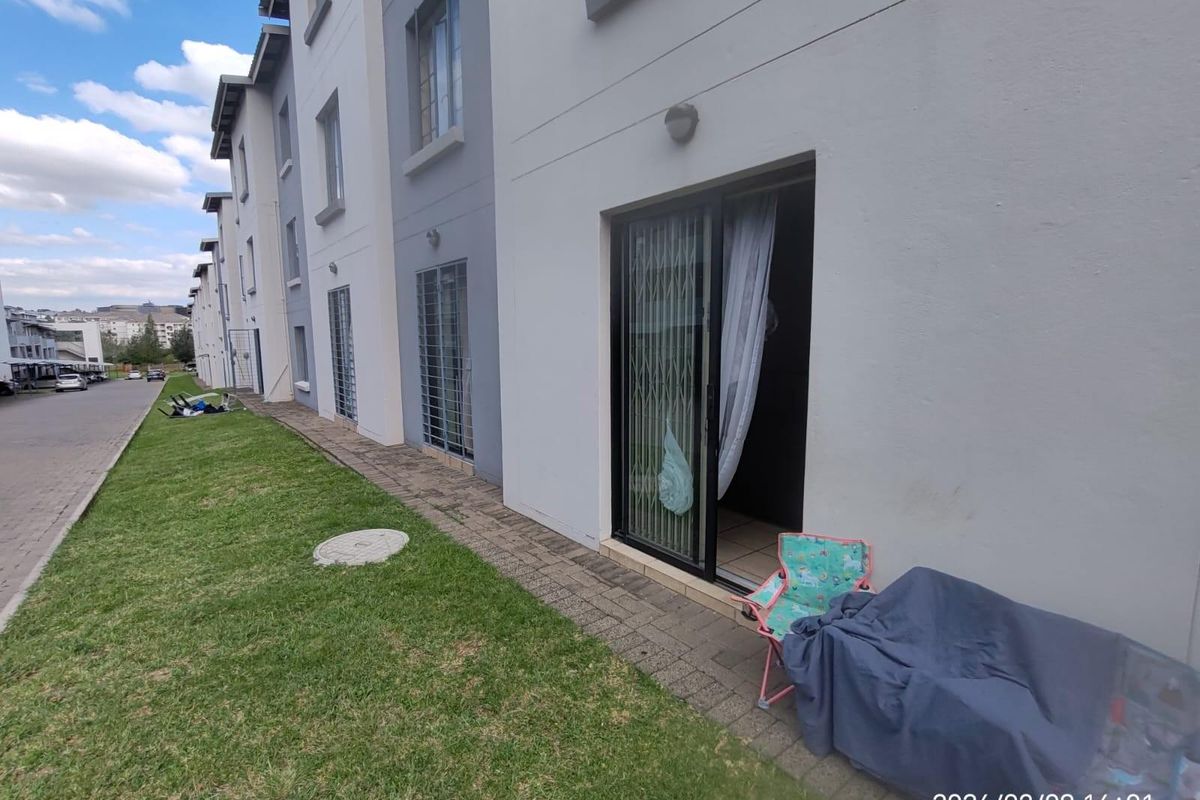 Exterior view of the apartment building showing ground floor access with sliding doors, security gates, and a paved walkway adjacent to a communal lawn.