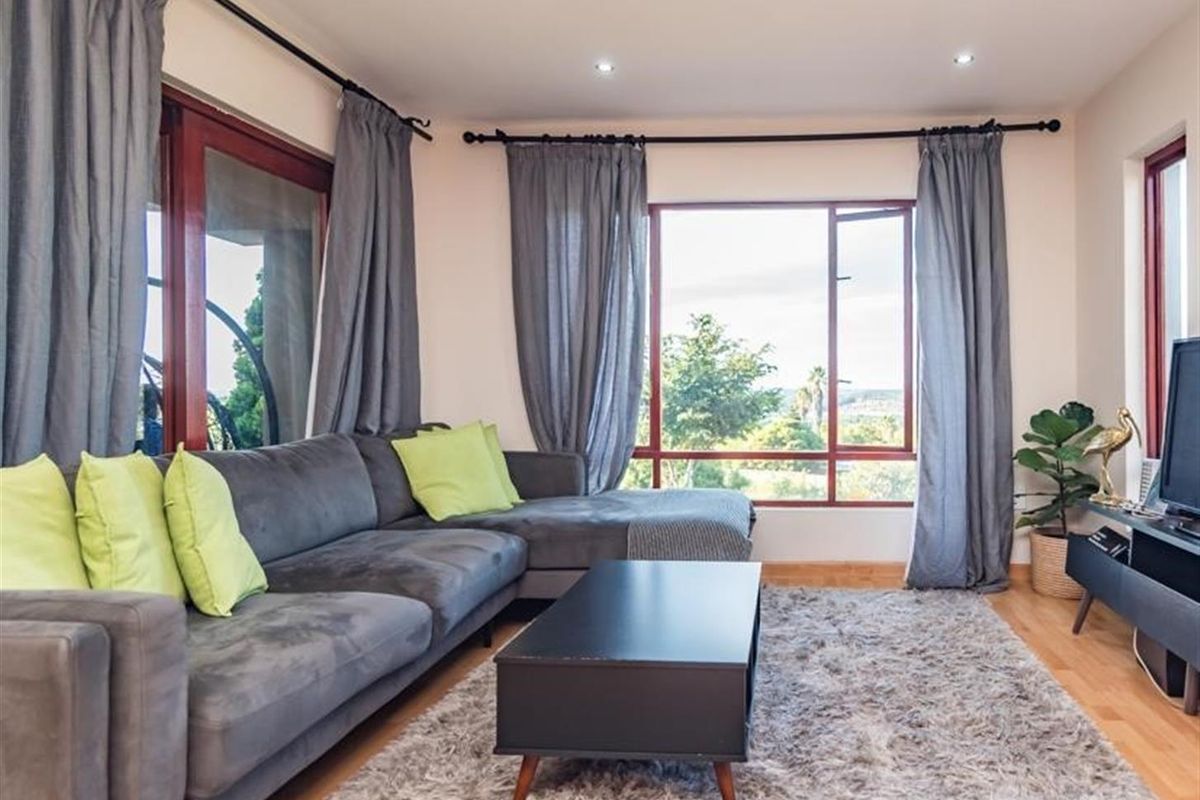 A bright and comfortable living room featuring large windows with views, recessed ceiling lighting, and warm-toned wooden flooring.