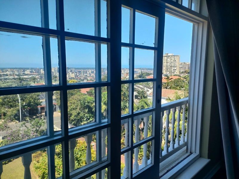 A view from an interior room looking out through classic timber-framed glass doors onto a balcony with an expansive urban and coastal cityscape.